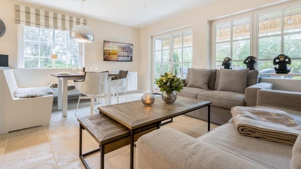 A spacious living room with a sofa, a coffee table, and a dining area with chairs and a table. There are large windows providing natural light and views of greenery outside. - House Süderhof, Seagull Whg. 13 - House Süderhof (Sylt-Ost)