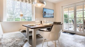 A dining area with a table set for two, a sofa, and a large window with a view of trees. - House Süderhof, Seagull Whg. 13 - House Süderhof (Sylt-Ost)