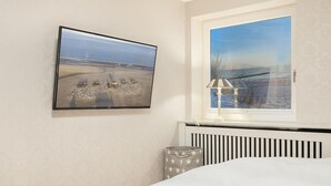 A bedroom with a bed, a window with a view of a body of water, a wall-mounted TV, and a bedside lamp. - House Süderhof, Seagull Whg. 13 - House Süderhof (Sylt-Ost)