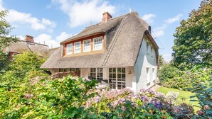 A traditional thatched-roof house with a white exterior, surrounded by lush greenery and flowering plants. - House Süderhof, Seagull Whg. 13 - House Süderhof (Sylt-Ost)