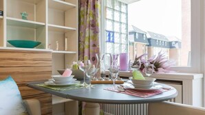 Ein Essbereich mit einem runden Tisch, gedeckt für vier Personen, umgeben von Stühlen. Regale mit Dekorationen und ein Fenster mit Blick auf Gebäude.