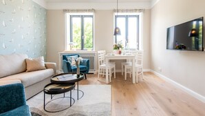 Una sala de estar moderna con un sofá, un sillón azul, una mesa de comedor con sillas, un televisor montado en la pared y una ventana con vistas a la vegetación.