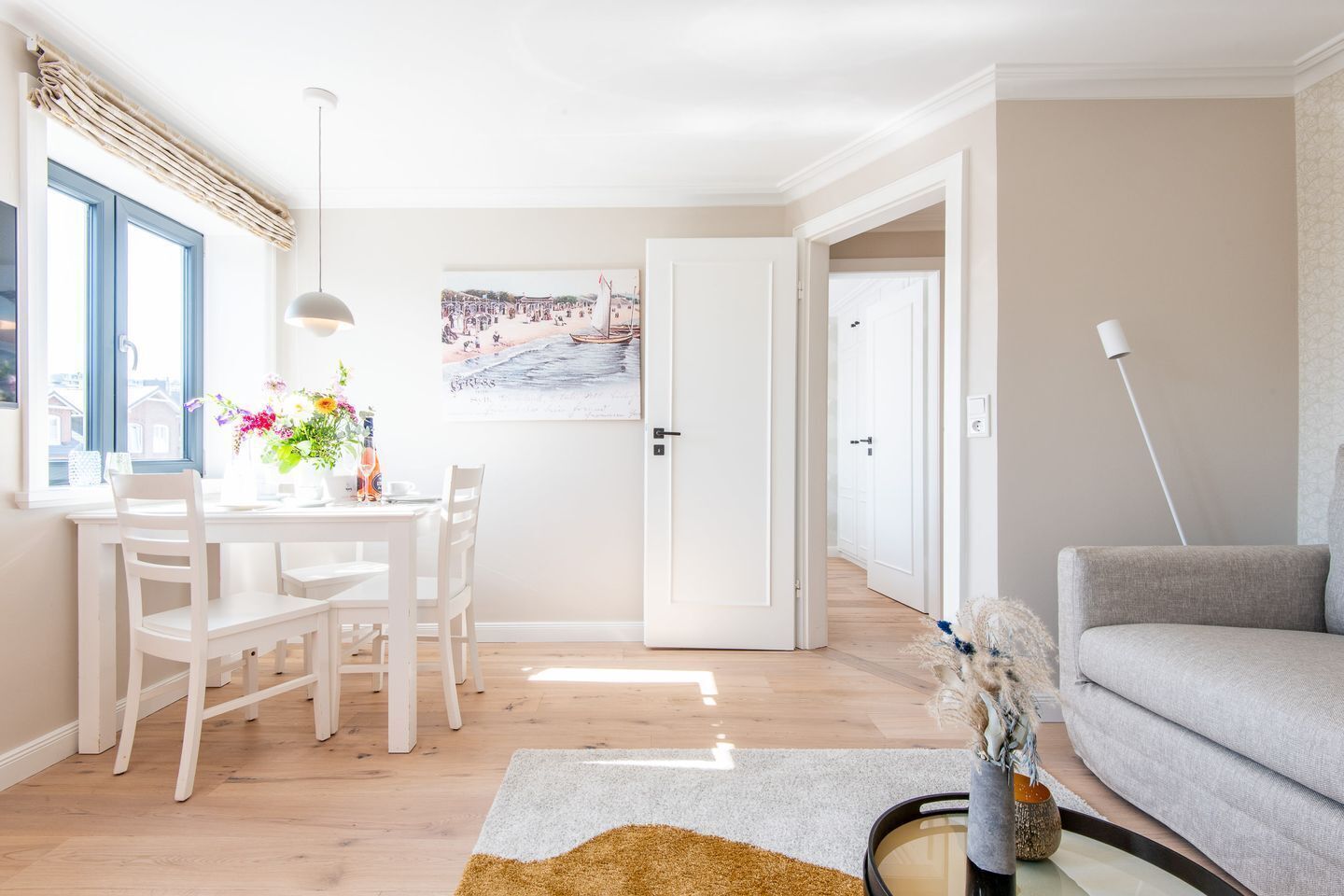 Ein helles Wohnzimmer mit Essbereich, einem grauen Sofa, einem weißen Tisch und einem Gemälde an der Wand.