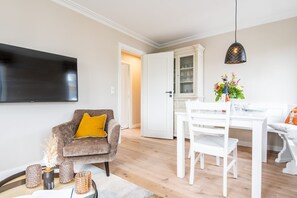 A modern living room with a flat-screen TV, a brown armchair with a yellow pillow, a white dining table with chairs, and a decorative vase with flowers.