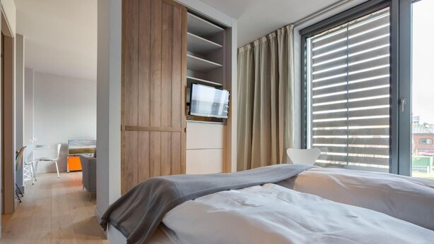 A modern hotel room with a bed, a TV, a desk, and a large window with blinds.