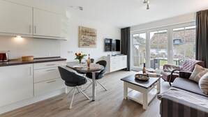 Une cuisine moderne avec des meubles blancs, un coin repas avec une table et des chaises, un salon avec un canapé et une table basse, et une grande porte-fenêtre coulissante donnant sur un patio.