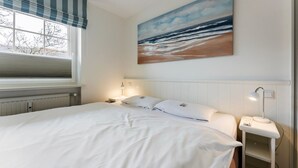 Une chambre d’hôtel avec un grand lit, une fenêtre avec un store à rayures, un tableau représentant une scène de plage, une table de chevet avec une lampe et une petite table de nuit avec un téléphone.
