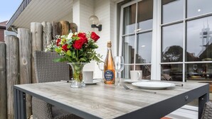 Une table à manger en plein air, avec une bouteille de vin, un vase de fleurs et une chaise en osier.