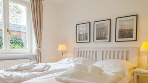 A neatly made bed with white linens, a bedside table with a lamp, and three framed pictures on the wall. - Gemütlich. Strandkorb. Ruhig. - Senwai-hüs, Whg. 2 "keitum" - Objekt-id: Hsw/02 - Senwai-hüs (Sylt)