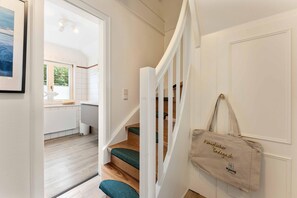 A staircase with a green cushioned step, a beige tote bag hanging on the wall, and a view into a kitchen area with white cabinets and a window. - Ruhe. Balkon. Gemütlich. - Senwai-hüs, Whg. 3 "morsum" - Objekt-id: Hsw/03 - Senwai-hüs (Sylt)