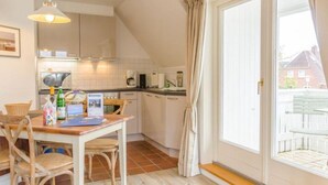 A kitchen with a dining table and chairs, a hanging light fixture, and a view of the outside through a sliding glass door. - Ruhe. Balkon. Gemütlich. - Senwai-hüs, Whg. 3 "morsum" - Objekt-id: Hsw/03 - Senwai-hüs (Sylt)