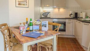 A compact kitchen with a dining table, chairs, a microwave, oven, and a coffee maker. - Ruhe. Balkon. Gemütlich. - Senwai-hüs, Whg. 3 "morsum" - Objekt-id: Hsw/03 - Senwai-hüs (Sylt)