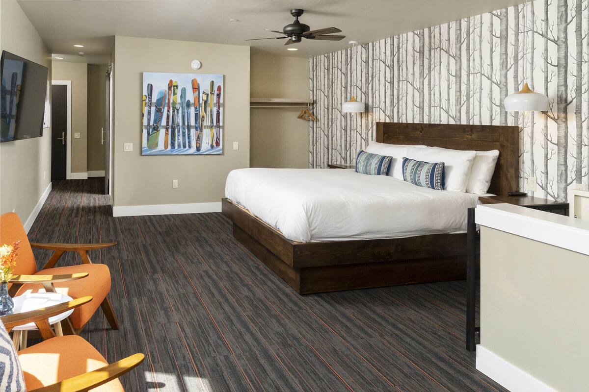 A hotel room with a large bed, a ceiling fan, a painting of skis, a wooden headboard, and a patterned wallpaper.