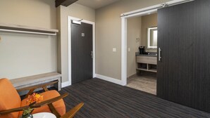 Habitación de hotel con alfombra oscura, un banco de madera, una mesa con un jarrón de flores y una puerta corredera que da a un baño.