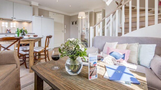 A modern living room with a wooden coffee table, a sofa with cushions, a dining area with a table and chairs, and a staircase. - Exquisit. Wellness. Modern. - Objekt-id: Hm/05 - Haus Magnus (Sylt)