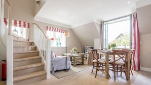 A spacious living area with a wooden staircase, a dining table with chairs, and a large window with curtains. - Exquisit. Wellness. Modern. - Objekt-id: Hm/05 - Haus Magnus (Sylt)