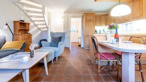 Una sala de estar moderna con un sofá, un sillón azul, una mesa de comedor con sillas y una zona de cocina con armarios de madera y una lámpara colgante.