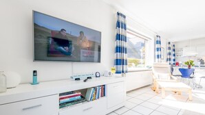 En moderne stue med et fladskærms-tv, et hvidt tv-møbel med bøger, et sofabord og et spiseområde med blå stole.
