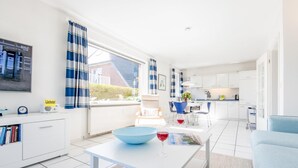 En moderne stue med en hvid sofa, et spisebord med en blå skål og vinglas, samt et køkkenområde med hvide skabe og blå stole.