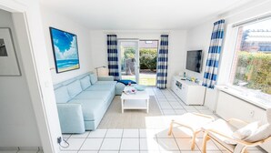 En moderne stue med en lyseblå sofa, et sofabord og et fjernsyn. Der er en balkon med udsigt over grøn bevoksning.