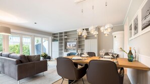 A modern living room with a sofa, a dining table set for four, and a kitchen area with a TV and shelves.