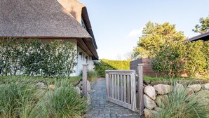 A pathway leading to a building with a thatched roof, surrounded by greenery and a wooden gate.