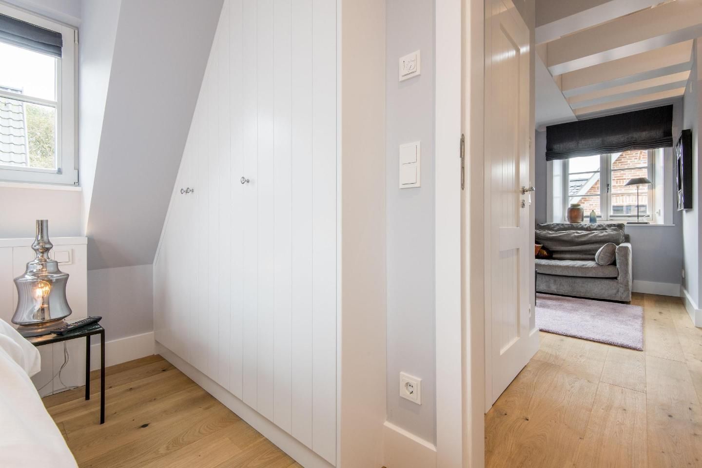 Een smalle gang met een schuine kap, een bed met een lamp en een bank in een hoek.