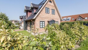 Uma casa de dois andares com telhado inclinado, fachada de tijolos e um pequeno jardim frontal com arbustos.