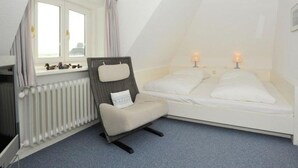 Un dormitorio bajo las tejas, pequeño y triangular, con una cama individual, una silla y una ventana.