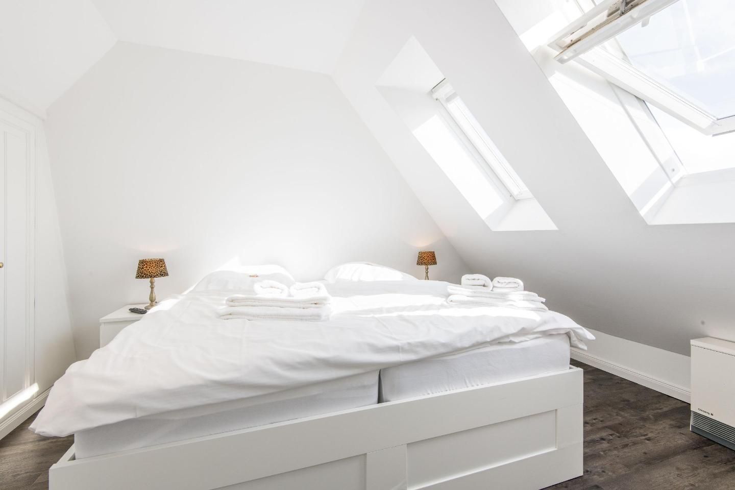Et lyst, moderne soveværelse med ovenlys, en seng med hvide sengetøj og et natbord med en lampe.