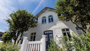 Un bâtiment à deux étages avec une façade blanche, une porte d’entrée sombre et une clôture blanche à piquets.