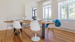 Un espace repas avec une table en bois, des chaises blanches et une vue sur l’extérieur grâce à de grandes fenêtres.
