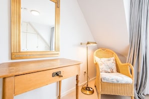 Ein Zimmer mit einem Holzkasten, einem Korbstuhl, einer Stehlampe und einem großen Spiegel.