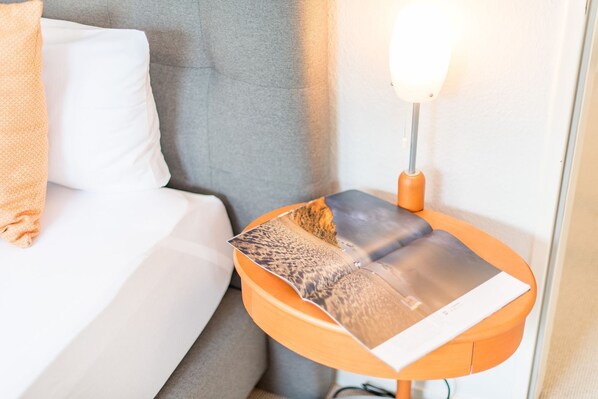A neatly made bed with a white pillow and a folded magazine on an orange side table next to it.