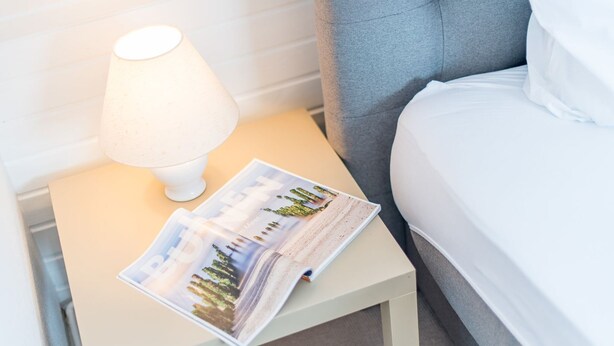 Ein ordentlich bezogenes Bett mit weißen Leinen, ein grauer möbelbezogener Kopfteil, ein kleiner Nachttisch mit einer weißen Lampe und eine Zeitschrift auf dem Tisch.