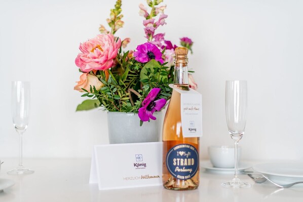 A bottle of I Heart Strand Perling champagne, a bouquet of flowers, and a welcome card with the König apartment logo. - Haus Dünenburg, Whg. 24 - Gemütlich. Zentral. Strandnah. - Objekt-id: Hdb/24 (Sylt)