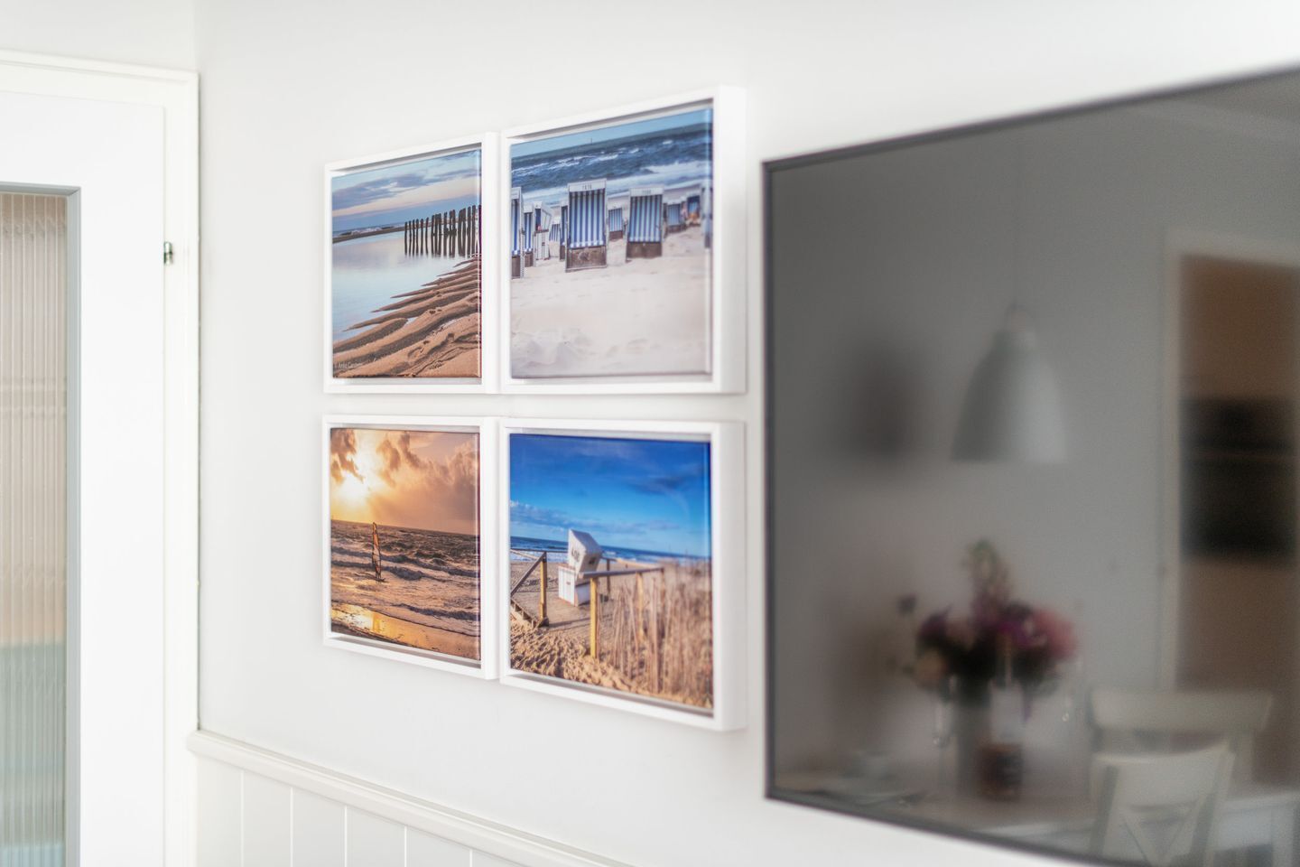 A wall with four framed beach scenes, a white door, and a window with a view of a beach and a chair.