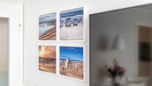 A wall with four framed beach scenes, a white door, and a window with a view of a beach and a chair. - Haus Dünenburg, Whg. 24 - Gemütlich. Zentral. Strandnah. - Objekt-id: Hdb/24 (Sylt)