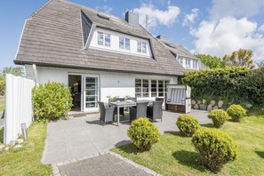 A two-story house with a patio, a dining area with outdoor furniture, and a garden with bushes.