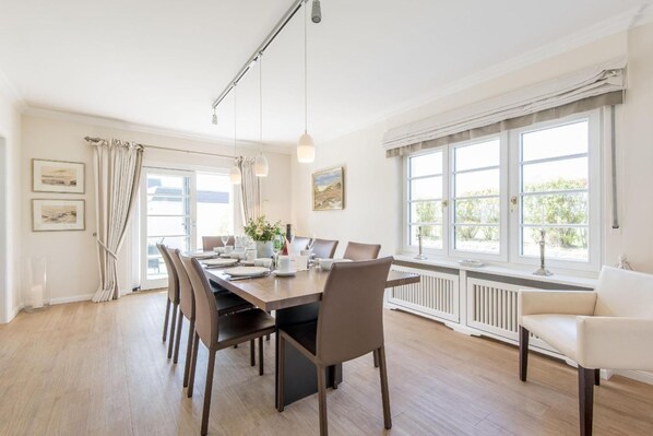 A dining room with a long table set for six, large windows with curtains, and a white radiator below.