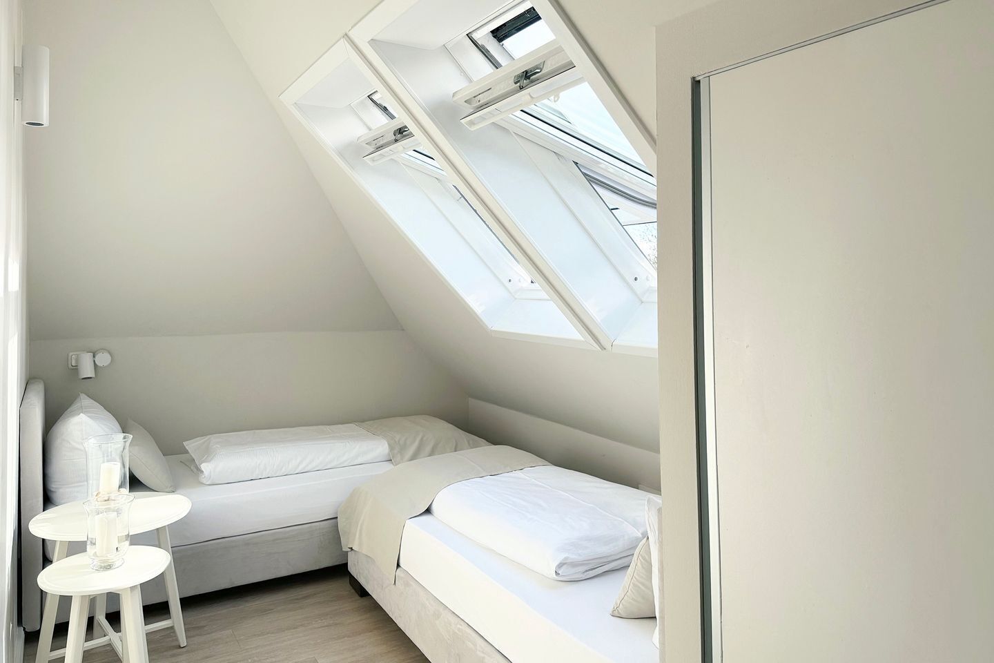 A small, white, sloped-roof bedroom with two beds, a nightstand, and a table with a candle.