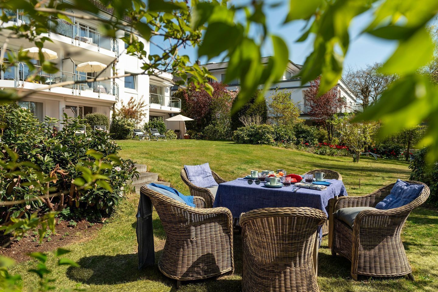 Eine Gartenpatio mit Korbmöbeln und einem Tisch, der für eine Mahlzeit gedeckt ist, umgeben von üppigem Grün und einem Gebäude im Hintergrund.