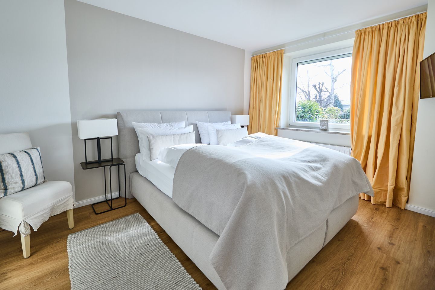 Ein modernes Schlafzimmer mit einem großen Bett, einem Nachttisch, einer Lampe und Blick ins Freie durch ein Fenster mit Vorhängen.
