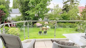 A balcony with wicker furniture, a glass railing, and a view of a well-maintained garden with a lawn, chairs, and a table. - Experience relaxation at TECHTs Apartmenthaus StrandPerle on Timmendorfer Strand (Timmendorfer Strand)