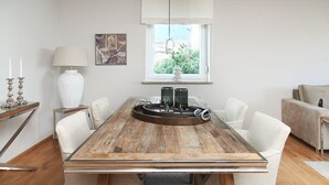 Ein Essbereich mit einem Holztisch, weißen Stühlen, einer Pflanzvase und einem Bild an der Wand.