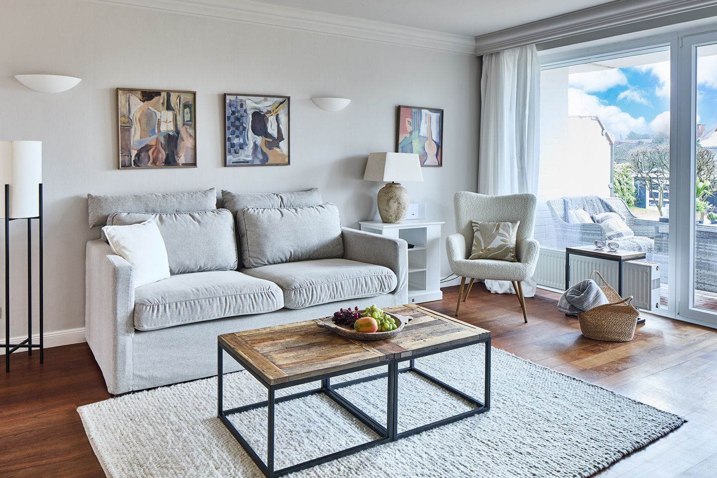 Ein modernes Wohnzimmer mit einem grauen Sofa, einem Holztisch und einem weißen Sessel. An der Wand befinden sich abstrakte Gemälde und ein großes Fenster mit Blick ins Freie.