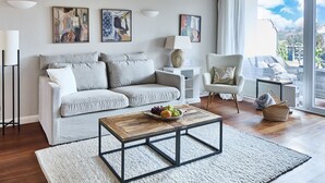 A modern living room with a grey sofa, a wooden coffee table, and a white armchair. There are abstract paintings on the wall and a large window with a view of the outdoors. - Welcome to TECHTS - TECHTs Apartmenthaus (Timmendorfer Strand)