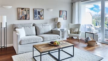 Ein modernes Wohnzimmer mit einem grauen Sofa, einem Holztisch und einem weißen Sessel. An der Wand befinden sich abstrakte Gemälde und ein großes Fenster mit Blick ins Freie.