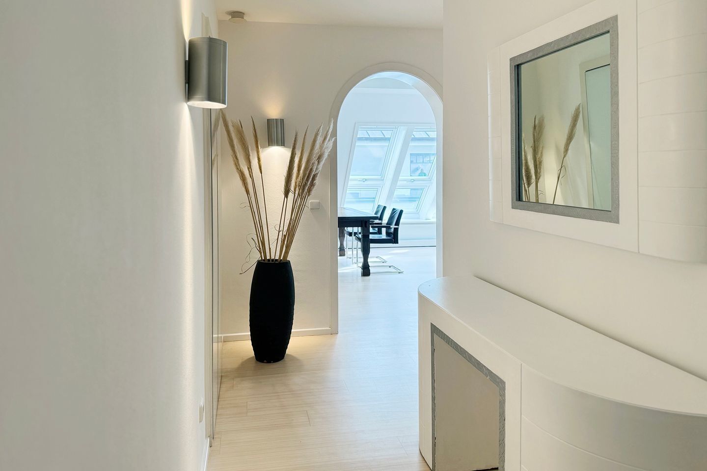 Un couloir moderne agrémenté d’un vase noir contenant des plumes d’herbe des pampas, un coin repas avec une table et des chaises, et une grande fenêtre laissant entrer la lumière naturelle.