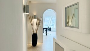 A modern hallway with a black vase of pampas grass, a dining area with a table and chairs, and a large window allowing natural light.